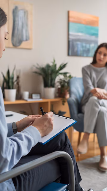 Sesión de terapia: primer plano de las manos de la terapeuta escribiendo en un portapapeles mientras, al fondo desenfocada, la paciente está sentada en un sillón