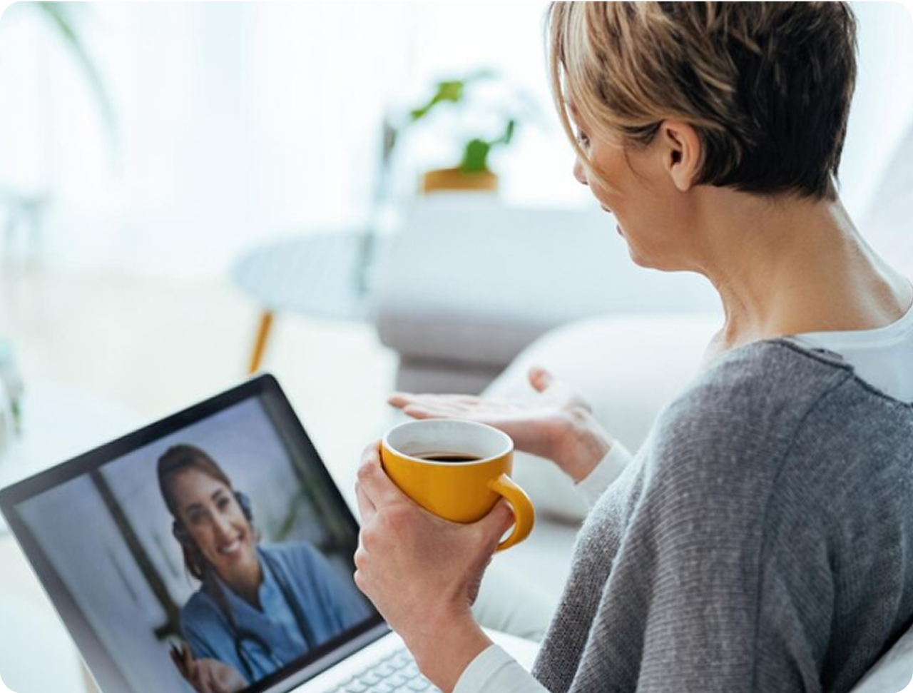 Mujer de perfil sostiene una taza amarilla mientras participa en una videollamada, con la imagen de la terapeuta visible en la pantalla del portátil.