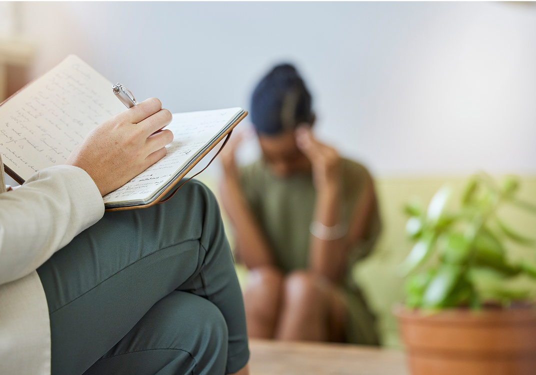 Mano de un terapeuta escribiendo en un cuaderno mientras al fondo, desenfocada, una persona cabizbaja se sujeta la cabeza con ambas manos.